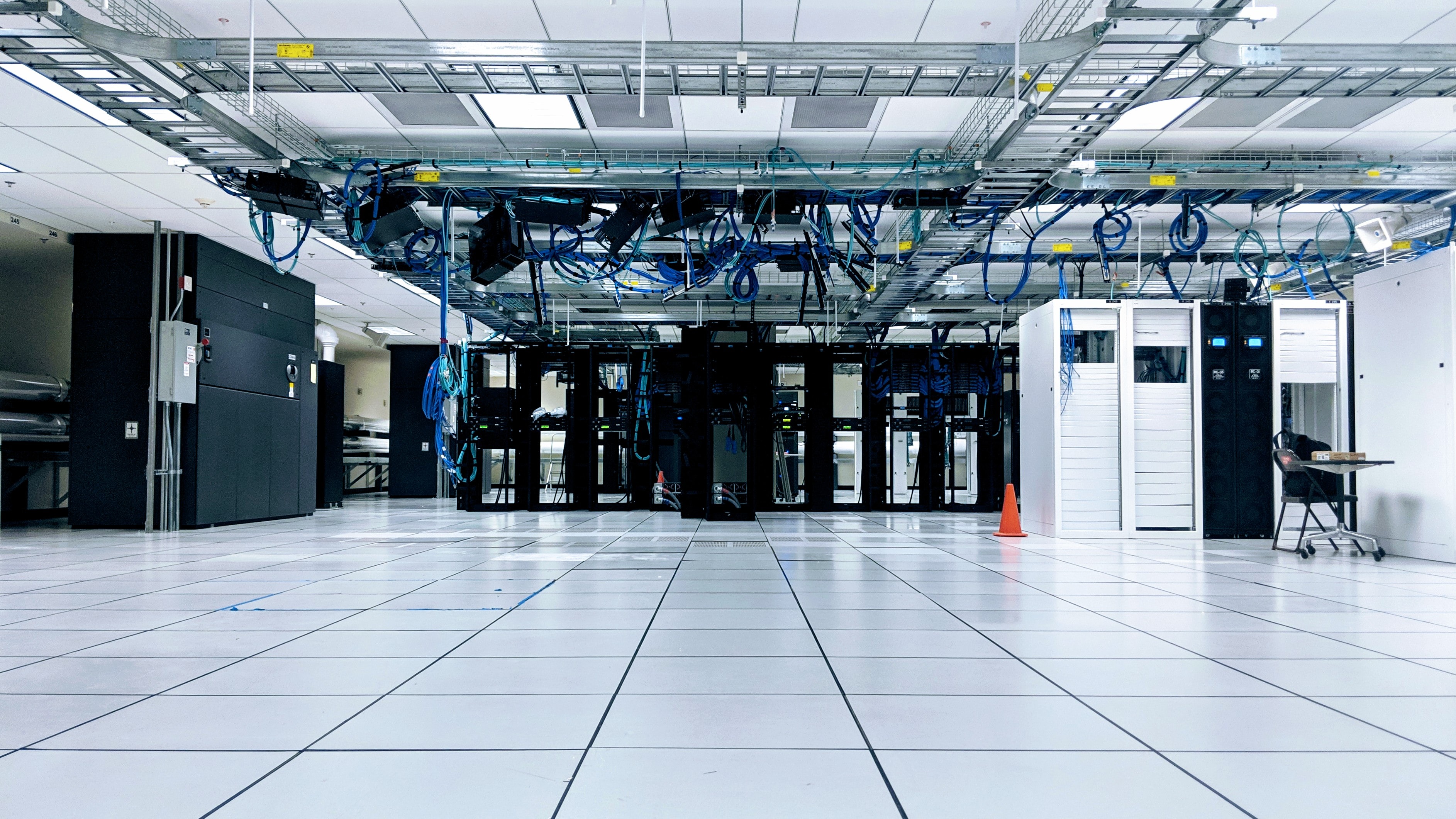 A bright, spacious data center with rows of black and white server cabinets, overhead cable trays filled with blue cables, and a white tiled raised floor in the foreground.